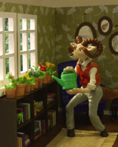 By Steph Brennan. Sculpture of creature that is a hybrid of a ram and fox. He is in his living room using a green watering can to water his plants.