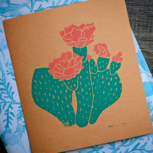 Linocut print of a flowering cactus. The flower is a coral pink colour and the print is done on orange paper.