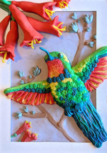 Brightly coloured hummingbird with vibrant, orange flowers made out of plasticine. Placed on a pale purple background.